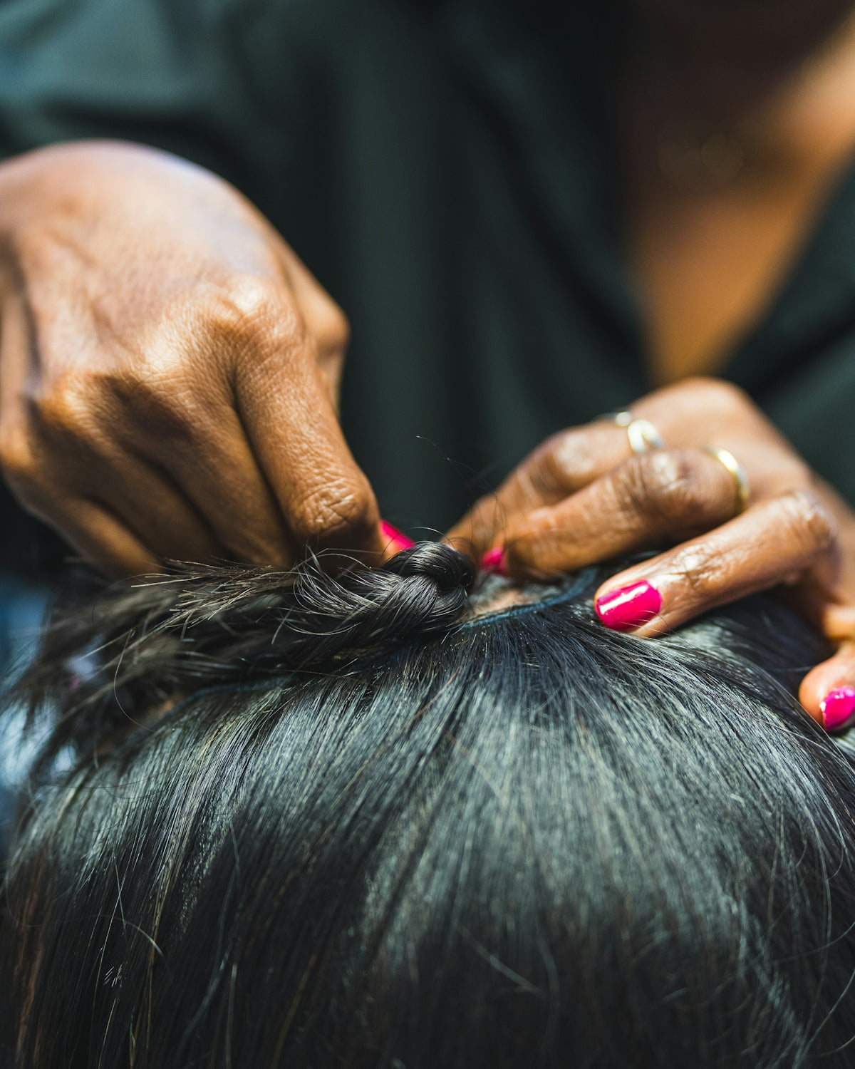 Hands braiding dark hair with pink nails.
