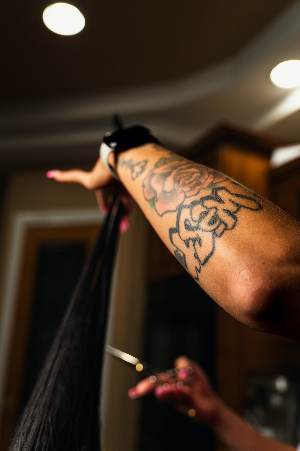 A tattooed arm holding a section of long dark hair with scissors